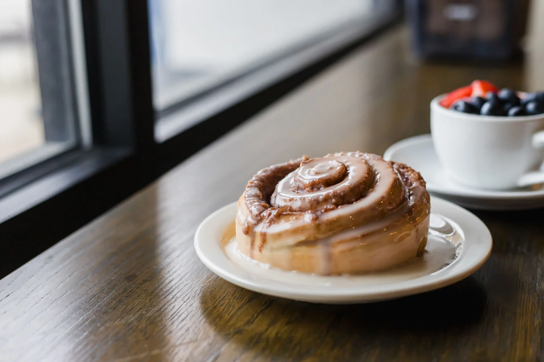 A glazed cinnamon roll on a plate by a sunlit window